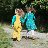Two children holding hands, walking on a path and wearing playful Ducky Zebra clothes. One in a pair of yellow dungarees, the other in a teal green dress with a highland cow print.