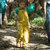 Girl wearing yellow cord dungarees, walking through a forest and holding onto her teddy. The dungarees have two big pockets and two playful appliqués of a highland cow and king fisher.