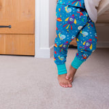 Photo of a child's legs, standing by the side of a bed. The child is wearing teal pyjamas with a fun, colourful space print and contrasting turquoise cuffs.