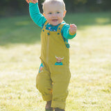 Toddler wearing mustard yellow dungarees with a highland cow peeking out of the pocket. They're walking across a field.