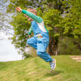 A boy leaping in the grass, wearing unisex dungarees by Ducky Zebra