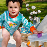 Image of a smiling baby playing outside, wearing a long sleeve turquoise stripe bodysuit with flame orange trim and roll-up arm cuffs. The bodysuit has two turquoise poppers on the neck and a sun, rainbow and cloud print on the chest