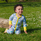 Image of a smiling baby wearing a long sleeve zip-up unisex baby romper with a light grey background and colourful repeat print pattern of a duck and zebra splashing in a puddle, riding a bicycle and running a three legged race. The images shows bright yellow cuffs, trim and a two way zip