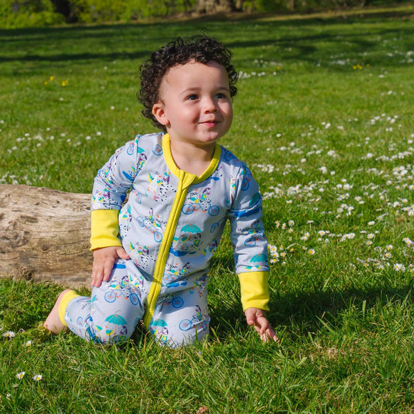 Image of a smiling baby wearing a long sleeve zip-up unisex baby romper with a light grey background and colourful repeat print pattern of a duck and zebra splashing in a puddle, riding a bicycle and running a three legged race. The images shows bright yellow cuffs, trim and a two way zip