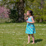 Happy girl blowing bubbles outside with bare feet and wearing a Ducky Zebra sleeveless summer dress with a turquoise stripe top half and a rainbow print bottom half. The picture shows one of the two deep pockets.