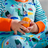 Close up image of a baby sitting down, wearing a Ducky Zebra colourful sleeveless romper with a turquoise background and repeat print pattern of a happy crocodile and elephant splashing at the seaside. The baby is holding an orange