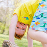 Image of a smiling, happy boy on all fours, balancing on a log, wearing a Ducky Zebra yellow t-shirt and seaside print shorts