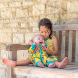 Image of a girl and baby sitting on a bench. The girl is cuddling the smiling baby. They are wearing Ducky Zebra twinning outfits. The girl is wearing a yellow dress with a repeat print of a crocodile and elephant enjoying a hot air balloon ride and playing together in the leaves. The baby is wearing a yellow sleepsuit with the same repeat print.