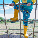 Child climbing up a rope net wearing Ducky Zebra teal trousers with a repeat print pattern of campervans and paddleboards. The trousers have a yellow trim around the pocket. The child is wearing yellow wellies.