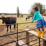 Image of a girl standing on a style by a field of cows. She's wearing a colourful Ducky Zebra skirt and hoody with a pair of wellies