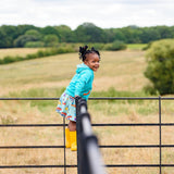 Image of a smiling girl standing on a fence wearing a colourful skort with a canoeing dog and hedgehog print with a turquoise hoodie and bright yellow wellies