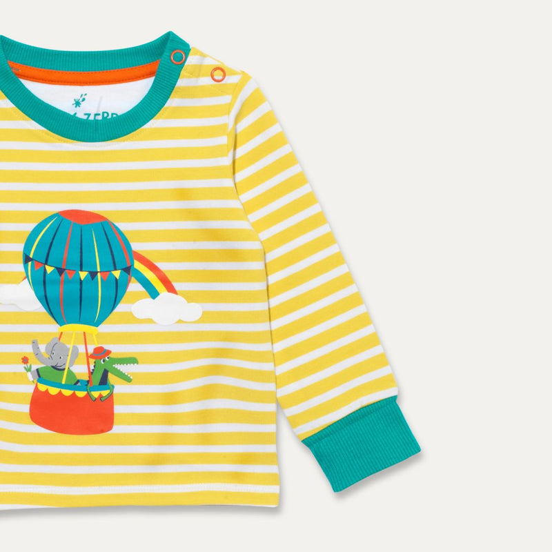 Partial image of a Ducky Zebra long-sleeve t-shirt. The t-shirt has yellow and white stripes with a print of a crocodile and elephant enjoying a hot air balloon ride together. There are two orange poppers on one of the shoulders. 