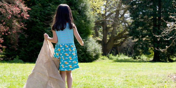 Image of the back of a girl wearing a Ducky Zebra sleeveless dress dragging a sack behind her, as she walks across the grass