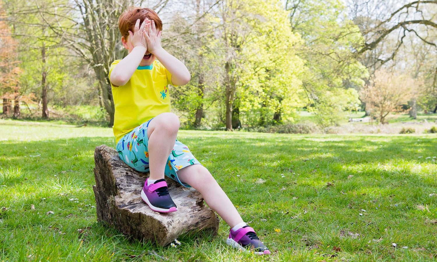 Image of a boy sitting on a log outside, covering his eyes with his hands. The boy is wearing a Ducky Zebra t-shirt, pair of shorts and pink trainers
