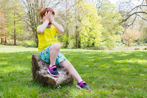 Image of a boy sitting on a log outside, covering his eyes with his hands. The boy is wearing a Ducky Zebra t-shirt, pair of shorts and pink trainers