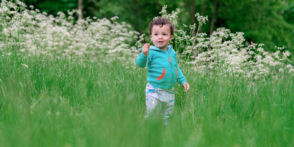 Image of a toddler amongst long grass, wearing a Ducky Zebra turquoise hoodie and Ducky Zebra joggers. The toddler is holding a dandelion. 