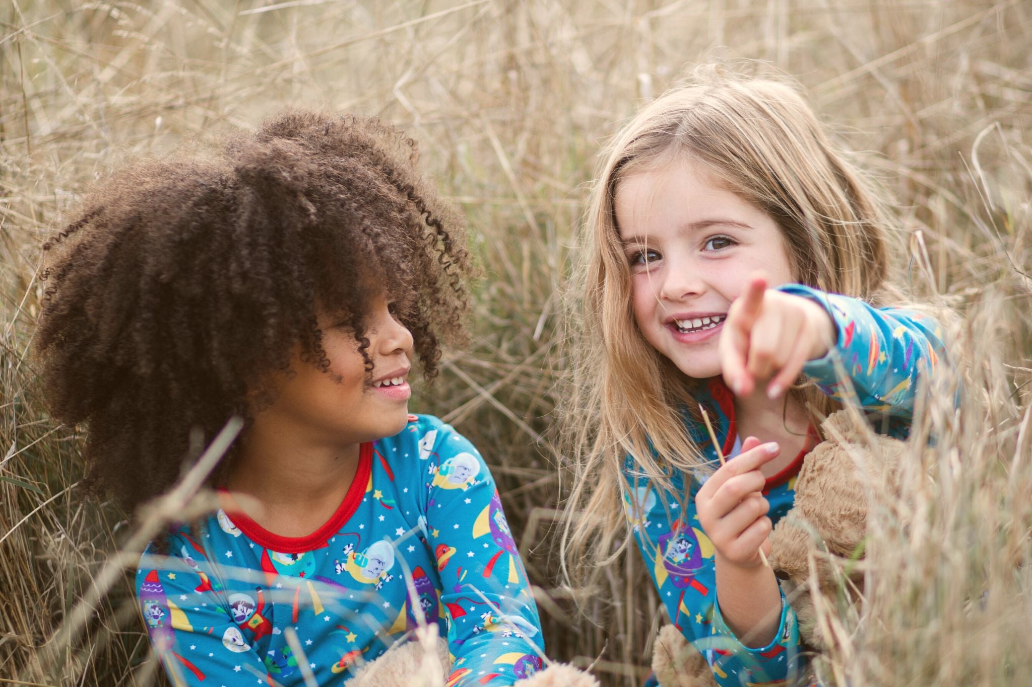 Image of two girls sitting in long grass pointing at the camera. They're both wearing Ducky Zebra space dresses