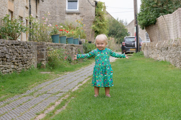 Smiling girl wearing a green dress with big pockets and a fun gardening print. The girl it's outside, smiling at the camera with her arms open wide.