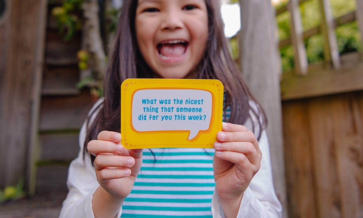 Smiling girl holding a conversation card saying 'What was the nicest thing that anyone did for you this week?' The girl is wearing a Ducky Zebra dress