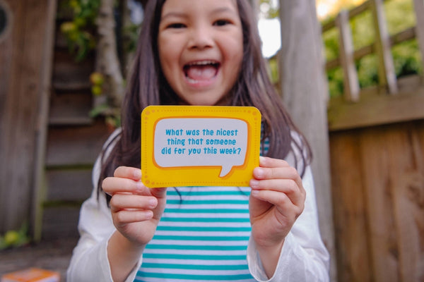 Smiling girl holding a conversation card saying 'What was the nicest thing that anyone did for you this week?' The girl is wearing a Ducky Zebra dress