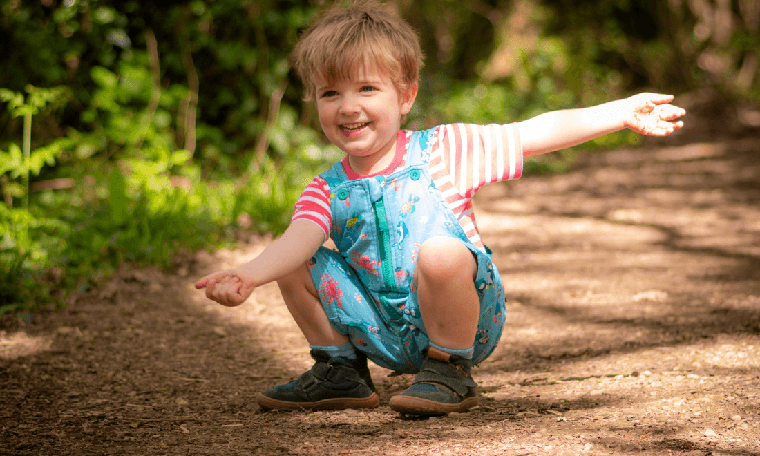 Boy crouching down outside, smiling at the camera, wearing Ducky Zebra short dungarees and a pink, striped T-shirt