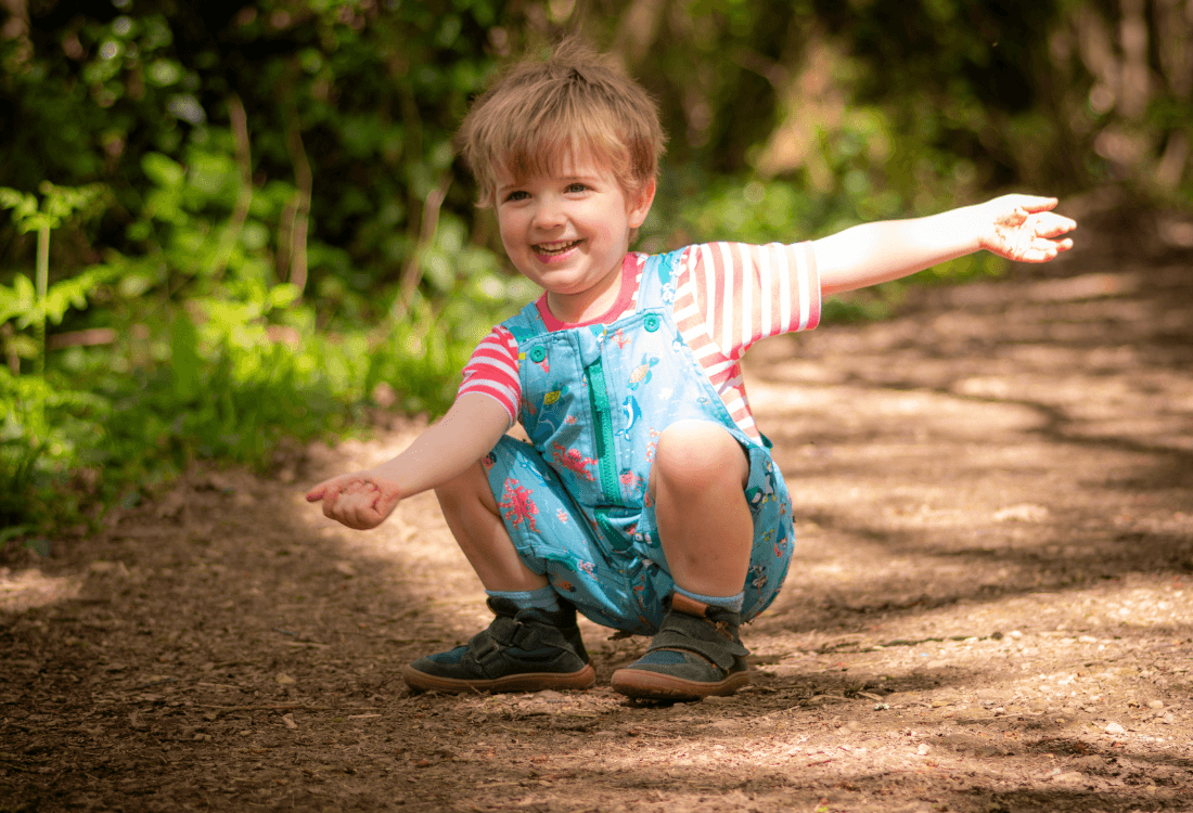 Boy crouching down outside, smiling at the camera, wearing Ducky Zebra short dungarees and a pink, striped T-shirt