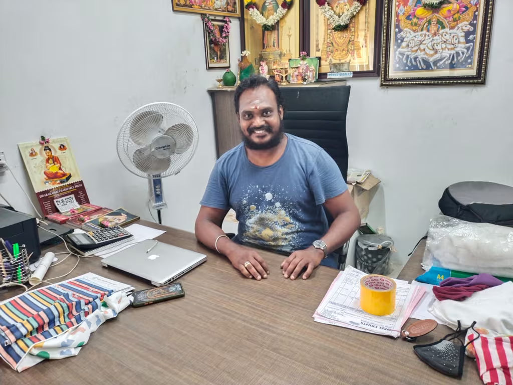 Man sitting at a desk with various items including a laptop, fan, and decorative items on the wall.