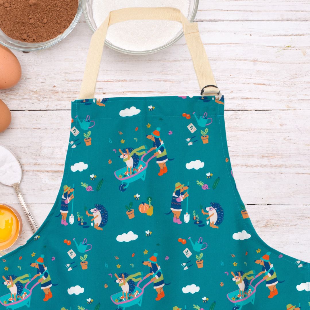 Close up of Ducky Zebra teal apron, with a repeat print pattern of dogs and hedgehogs playing in a wheelbarrow and tending to their vegetables. The apron is sitting alongside eggs, sugar and flour.