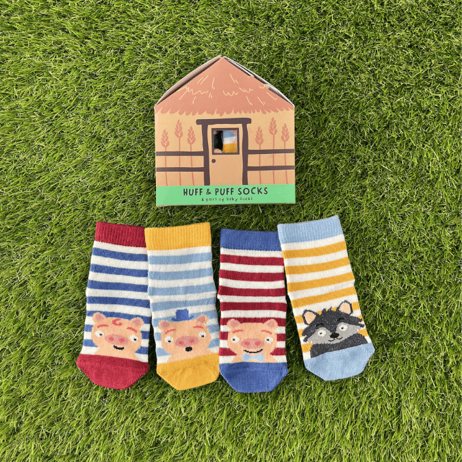 Four pairs of striped socks, three with pigs on and one with a wolf on - laid out in front of a gift box which says 'Huff and Puff Socks'