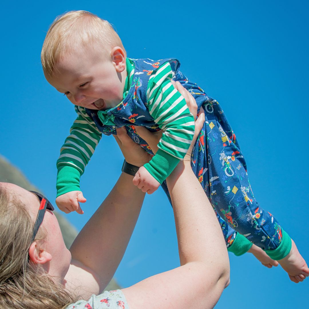 Smiling baby being thrown into the air, wearing a Ducky Zebra unisex baby outfit, including a green stripe top and blue dungarees