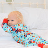 Image of baby girl lying in bed wearing a unisex, sustainable sleepsuit with light blue background and fun, farmyard print including tractors, pigs and sheep. The sleepsuit has contrasting red cuffs.