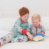 A baby and boy sitting together in bed wearing twinning Ducky Zebra pyjamas and sleepsuit with a fun tractor print and red cuffs. The boy is helping the baby read a book.