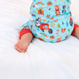 Image of a baby kneeling in bed wearing a unisex, sustainable sleepsuit with light blue background and fun, farmyard print including tractors, pigs and sheep. The sleepsuit has contrasting red cuffs.