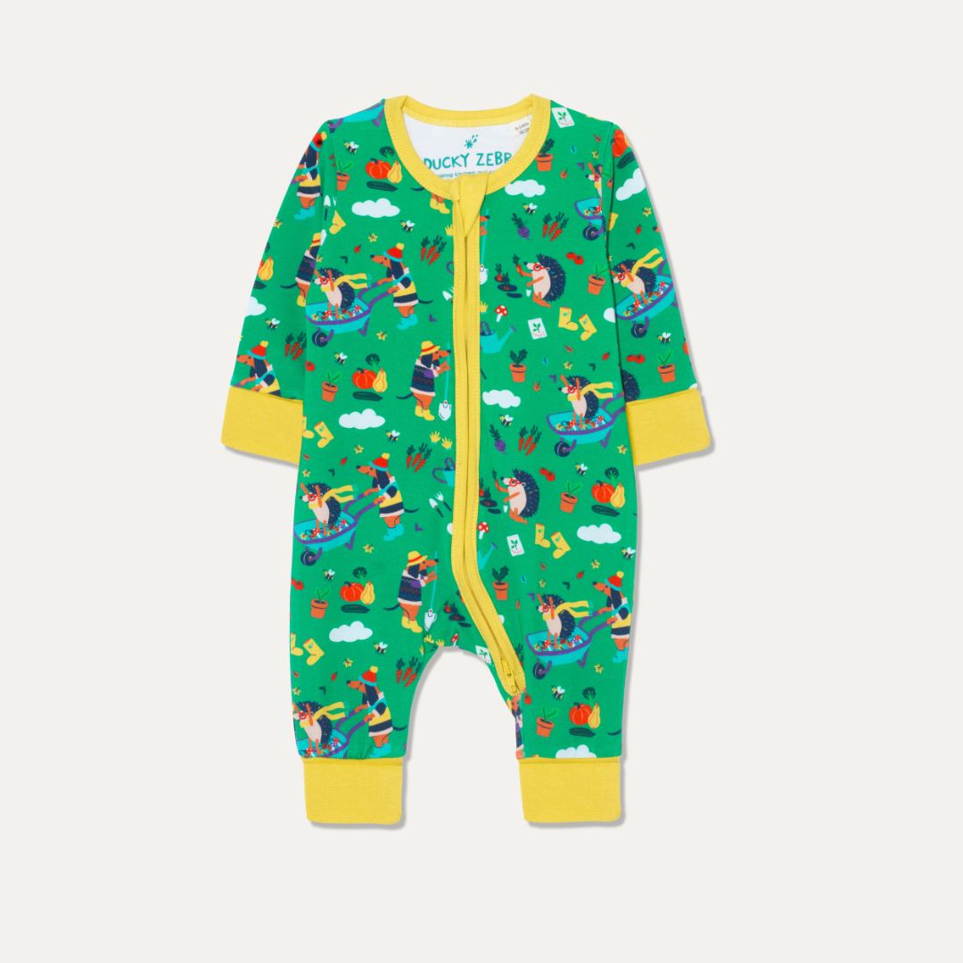 Baby zip-up sleepsuit with a green background and repeat print of a dog and hedgehog playing with a wheelbarrow and gardening together. The sleepsuit has a contrasting yellow zip, cuffs and chin guard.   
