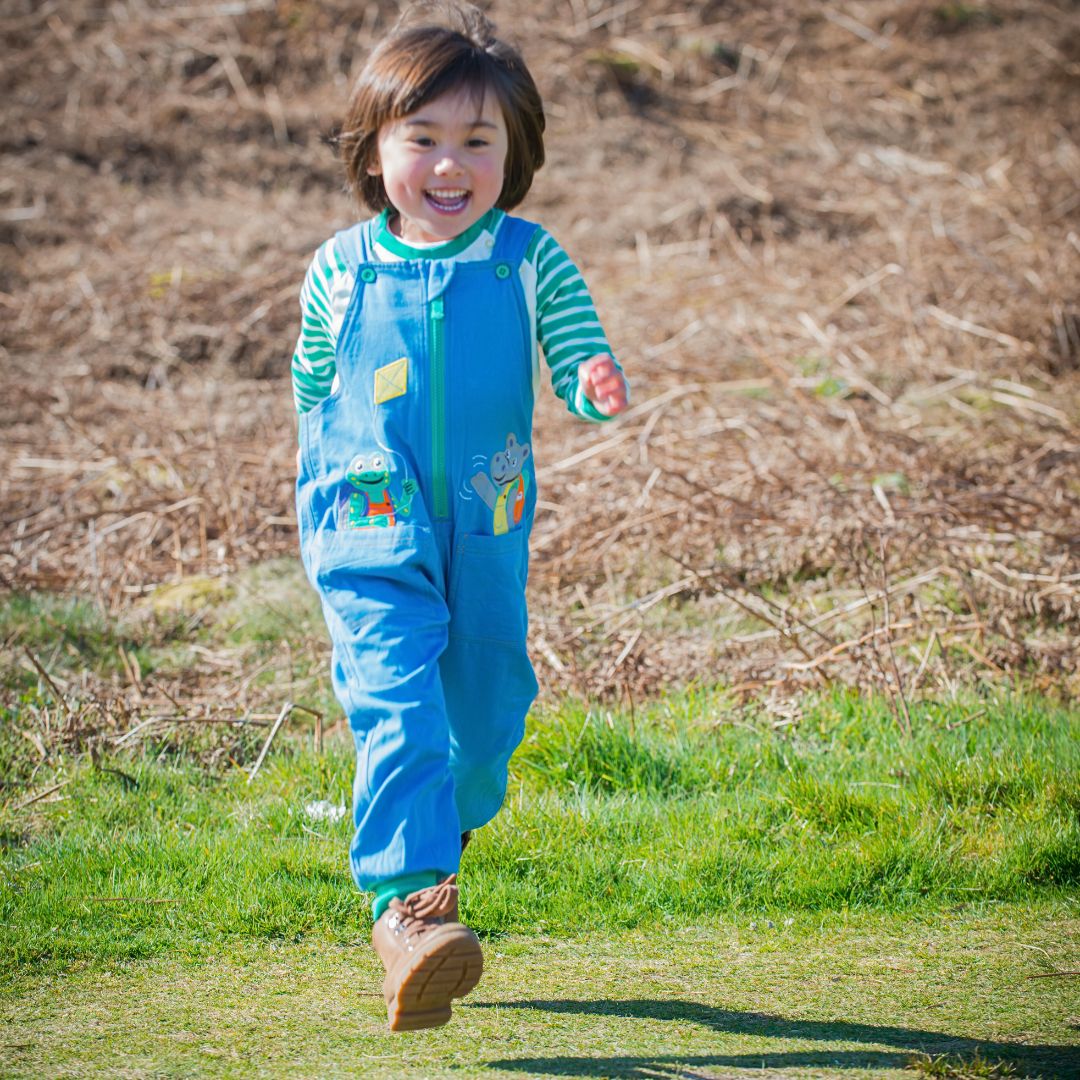 Girl running in blue organic cotton dungarees with a frog and hippo appliqué peaking out of two front pockets. The girl is running across the Yorkshire Moors with a big smile on her face