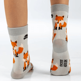 A pair of light grey children's unisex socks with an orange and white fox pattern, shown on a person's feet against a white background. The brand name 'HEDGY SOCKS' is printed vertically on the sole of one sock.