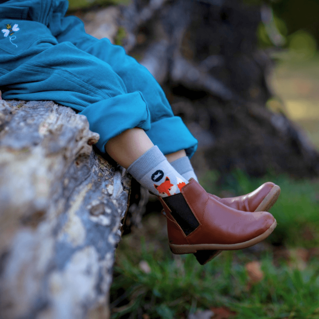 A child sitting outdoors on a log, wearing teal dungarees, brown ankle boots, and grey socks with a fox on it, which is peeking out of the boots.