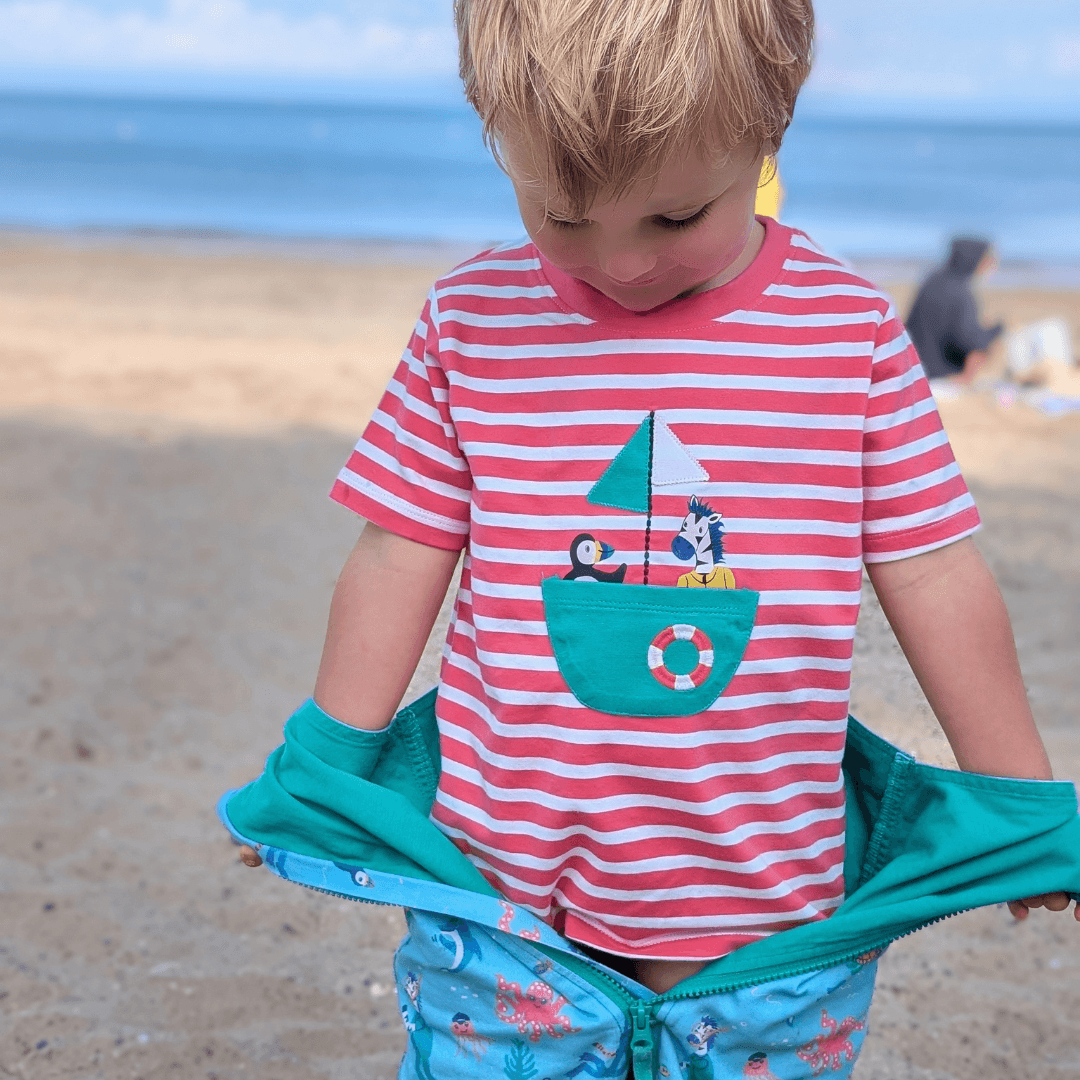 Toddler looking down at their coral-pink striped t-shirt with a puffin and zebra sailing in a boat shaped pocket The child is standing on the beach.