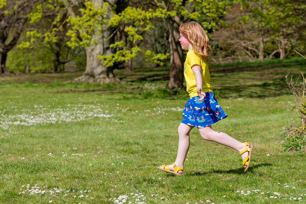 Child in a yellow shirt and blue skirt walking on a grassy field with trees in the background
