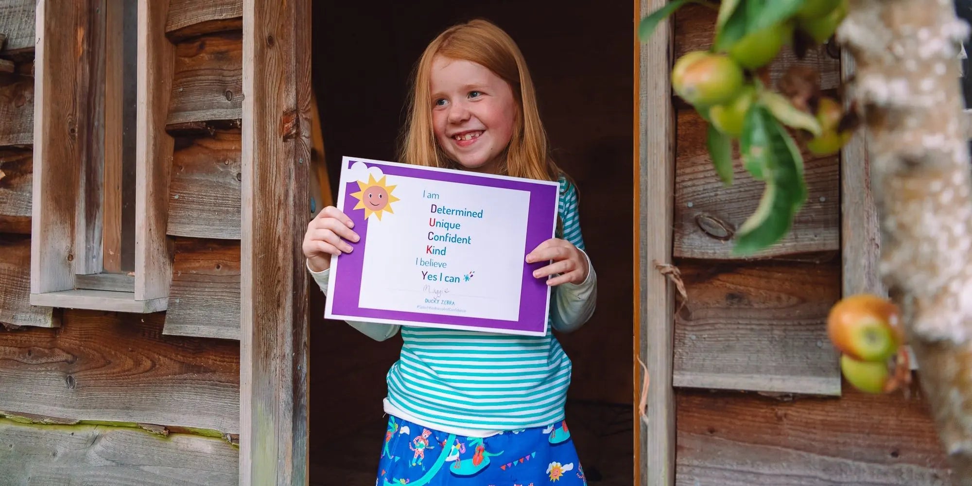 Young girl holding a purple sign with text, standing in front of a wooden door with apples on a tree.
