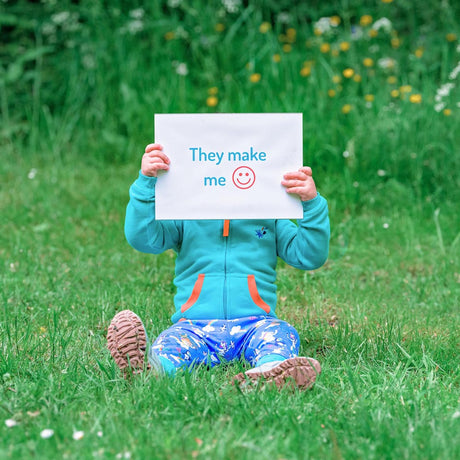 Image of toddler holding a sign in front of his face stating "They make me happy". The toddler is sitting in the grass, wearing Ducky Zebra clothes