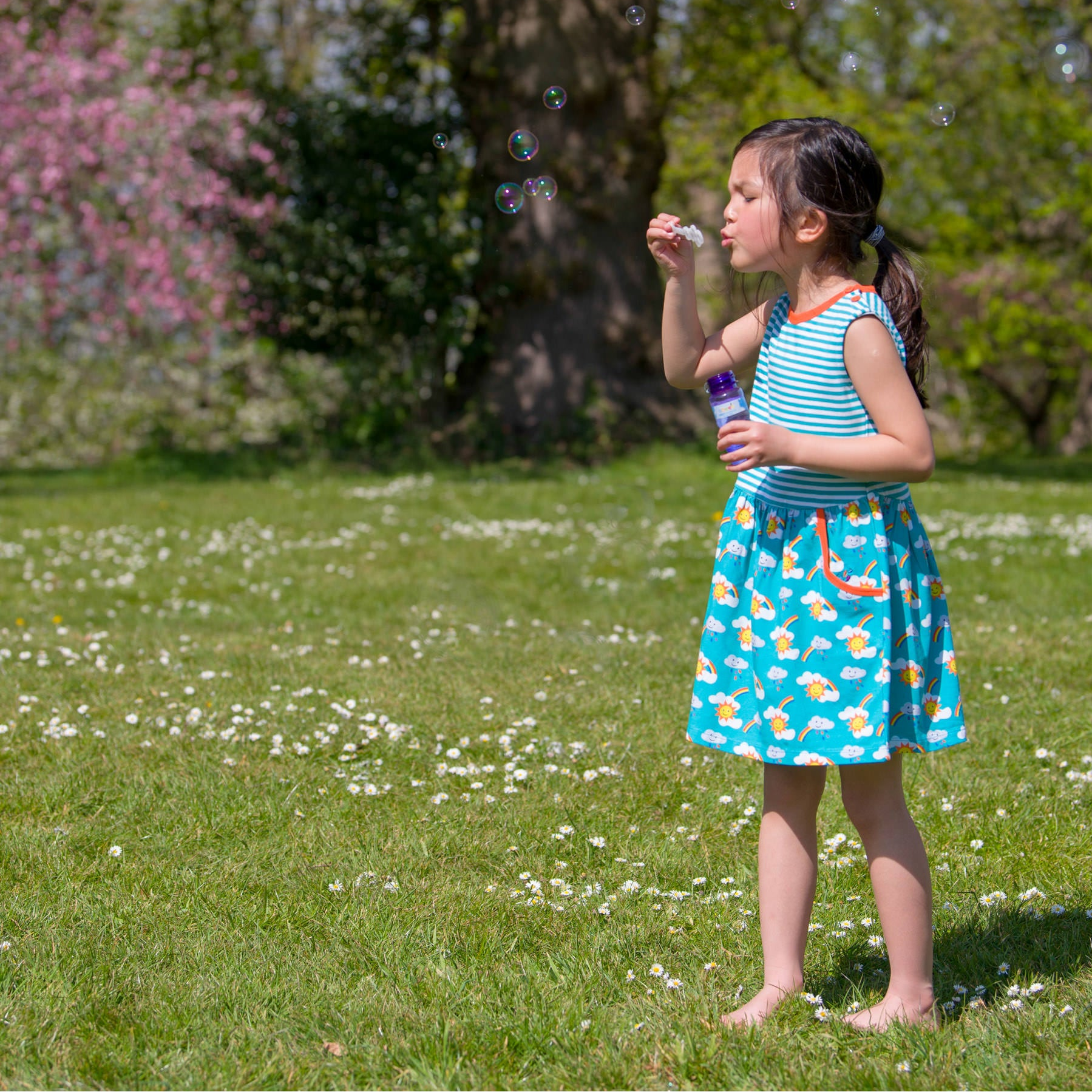Young girl blowing bubbles in a park with green grass and flowers.