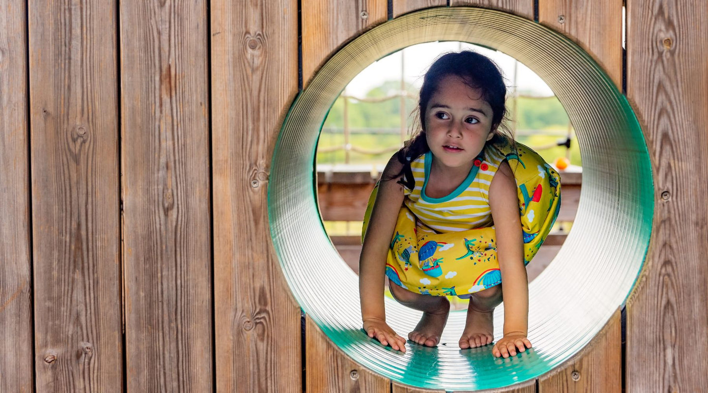 Girl peeking out of a tunnel wearing a sleeveless yellow Ducky Zebra dress with pockets and pictures of crocodiles and elephants in hot air balloons