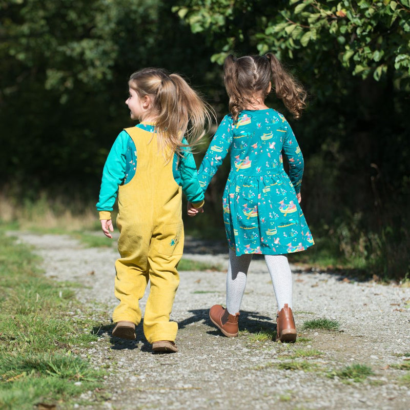 Two children holding hands, walking on a path and wearing playful Ducky Zebra clothes. One in a pair of yellow dungarees, the other in a teal green dress with a highland cow print. 