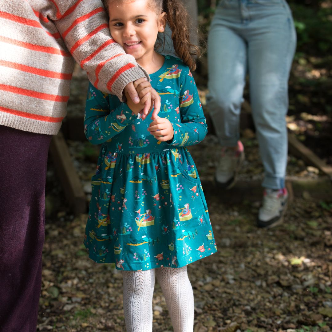 Girl wearing Ducky Zebra's 'Highland Pals' teal dress, holding onto her parent's hand as she walks through the woods