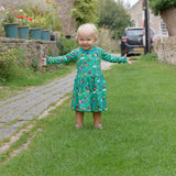 Smiling toddler outside wearing a green Ducky Zebra dress with a fun print of dogs and hedgehogs playing together. 
