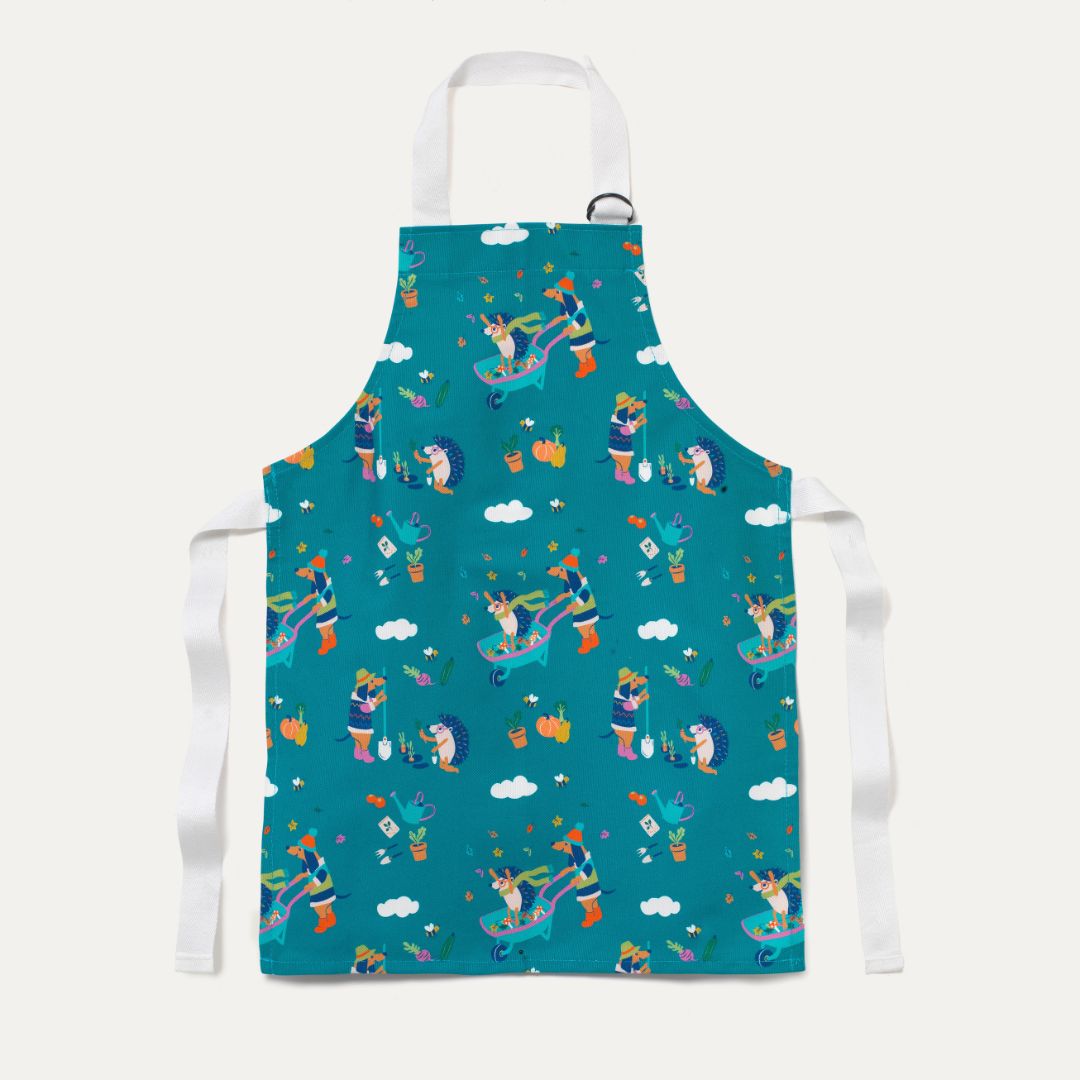 Kids Green Apron with fun dog and hedgehog repeat print. The dog and hedgehog are busy gardening and playing with a wheelbarrow in the leaves. Inspired by the Cotswolds.