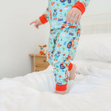 Image of a child standing on bed wearing Ducky Zebra 'farmyard fun' pyjamas. The pyjamas are light blue with red cuffs and a fun repeat print pattern, including pigs, sheep, tractors, bees and rainbows.