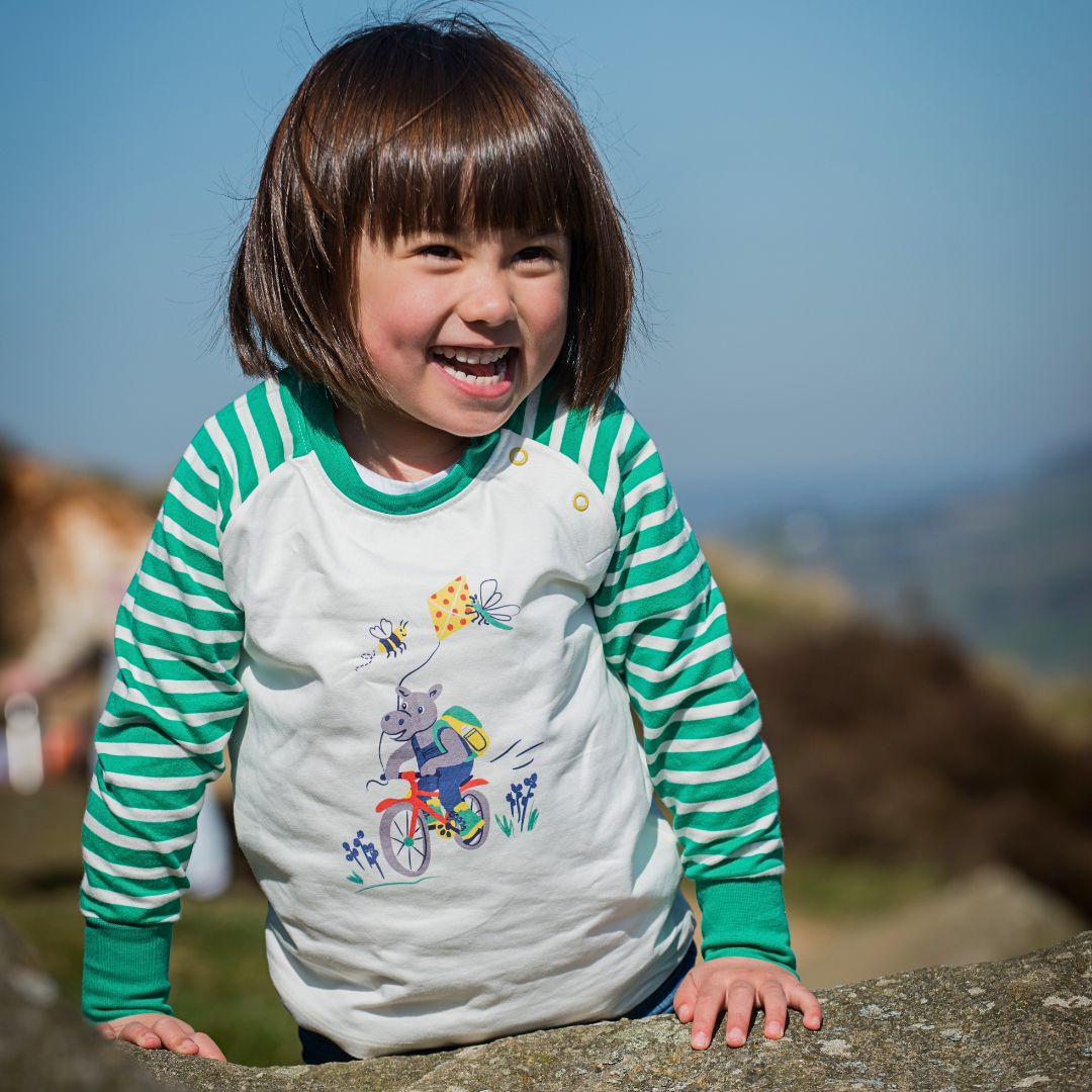 Smiling girl wearing a Ducky Zebra cream raglan top with a print of a cycling, kite flying hippo and a green and white stripe arm and cuff.