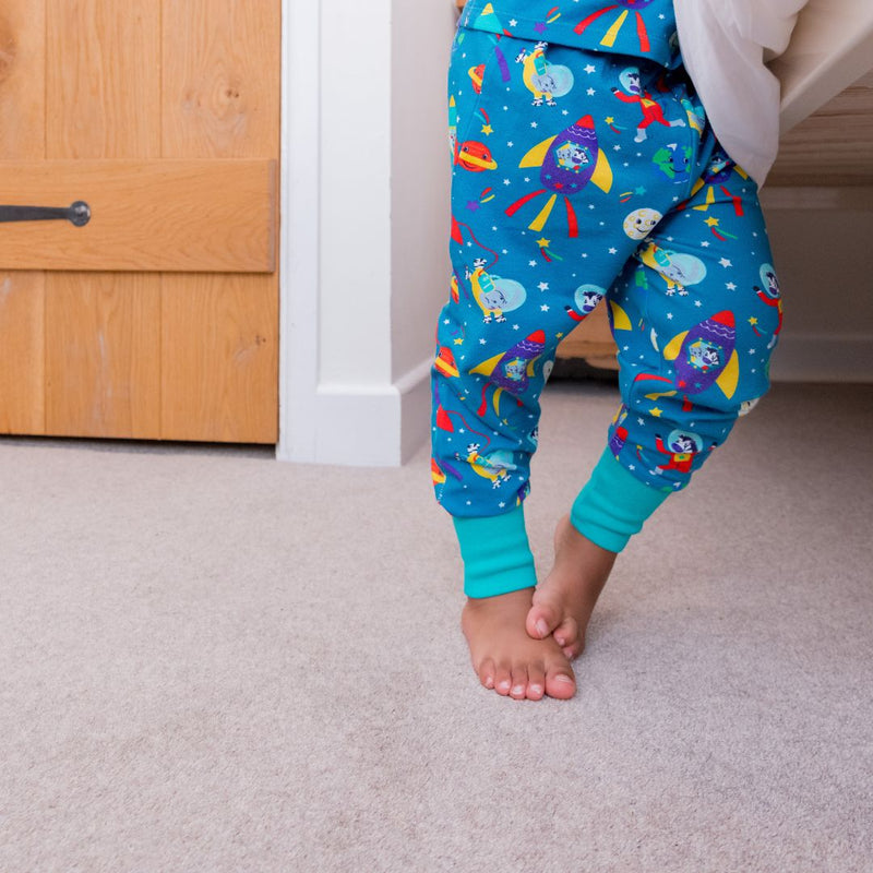 Photo of a child's legs, standing by the side of a bed. The child is wearing teal pyjamas with a fun, colourful space print and contrasting turquoise cuffs.